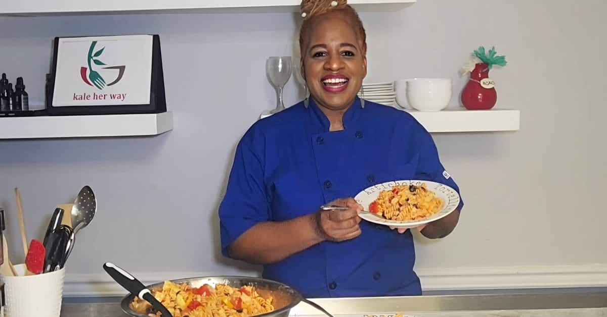 Raw Chef Gail cheerful in a blue uniform is smiling at the camera and holding a plate of pasta. She is standing in a kitchen with culinary tools organized on the counter and shelves behind her. There is a logo displayed on a digital frame that reads "kale her way." The overall environment suggests a cooking show or a culinary presentation setup.